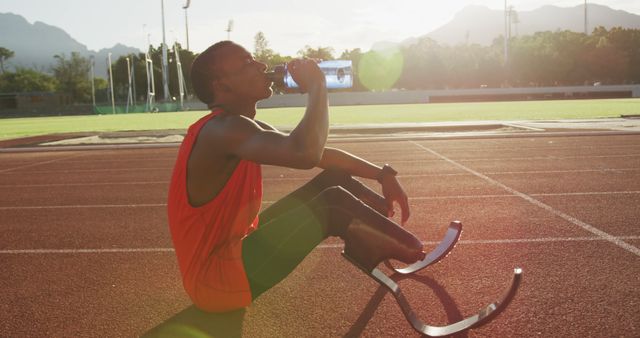 Paralympic athlete wearing sportswear is hydrating while taking a break on a track field during summer. He is seen drinking water while sitting, displaying prosthetic legs. Mountains and greenery are visible in the background, with the sun setting, providing a warm, inspiring ambiance. Ideal for topics on adaptive sports, determination, fitness motivation, and summer training.