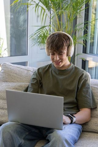 Teen male sitting on couch wearing headphones using laptop for remote learning and gaming