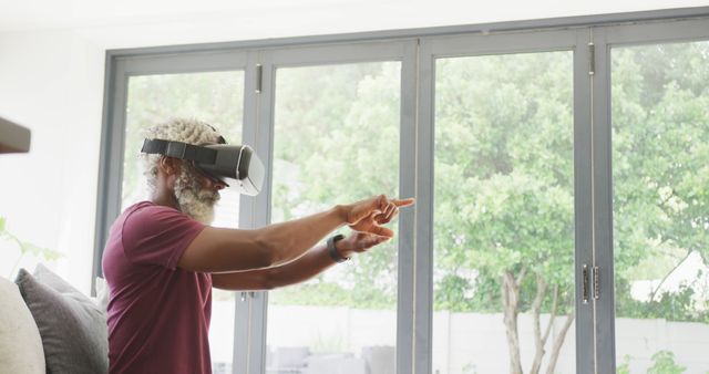 Middle-aged man using VR headset at home with large windows in the background. The man is enthusiastically interacting with the virtual environment, pointing at various objects. This depiction of someone fully engaged in a virtual reality experience highlights themes of technology, innovation, and modern entertainment. Suitable for promoting VR technology, gaming, tech products, and innovative home setups.