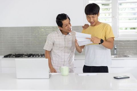 Asian father and teenage son reading letter together in bright modern kitchen