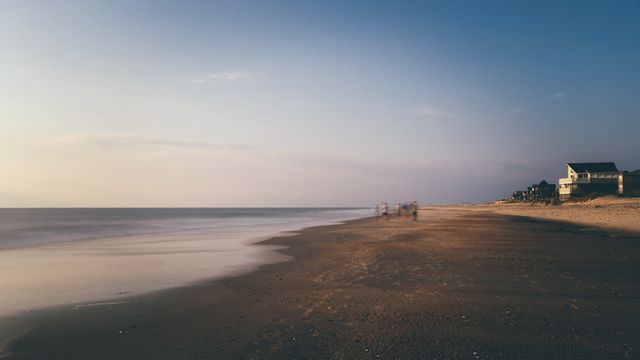 Wide-angle view of a serene beach at sunset showing calm ocean waves lapping against the sandy shore. In the distance, a few figures are walking near the water's edge, giving a sense of tranquility. A lone house stands by the coastline, hinting at an idyllic seaside living setting. This idyllic scene is perfect for travel blogs, tourism promotions, or nature-themed calendars, emphasizing peace and natural beauty.