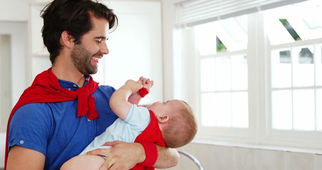 Dad wearing red cape holding baby dressed in blue in bright room. Perfect for parenting, family blogs, fatherhood articles, promoting positive family relationships, childcare advertisements.
