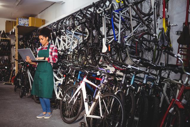 Mechanic standing in bike repair shop using a laptop surrounded by bicycles and tools. This image can be used to depict technology in traditional settings, small business operations, female professionals in technical roles, or modern repair and maintenance shops.