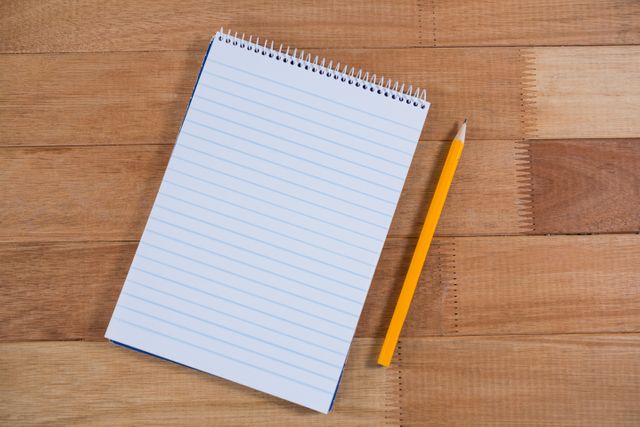 Close-up of a blank notepad and a yellow pencil on a wooden table. Ideal for illustrating concepts related to writing, note-taking, education, office work, and organization. Suitable for use in blogs, educational materials, office supply advertisements, and productivity articles.