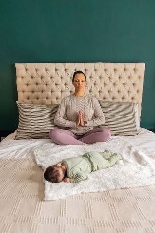 Mother meditating on bed while baby resting on blanket in cozy tufted green bedroom