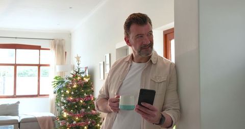 Mature man relaxing by christmas tree with smartphone and mug