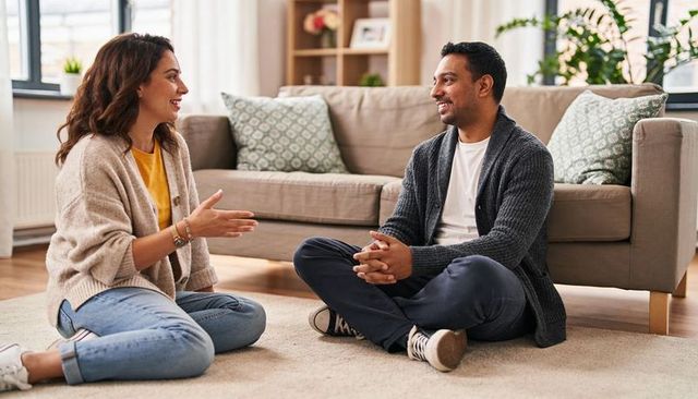 Couple having warm conversation sitting on living room rug wearing cardigans and jeans