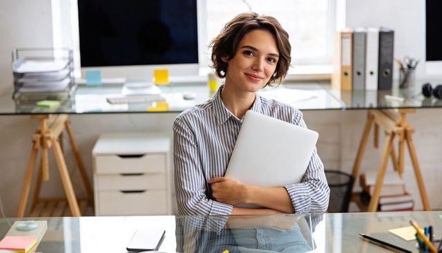 Smiling creative professional hugging laptop at glass desk in bright home office workspace