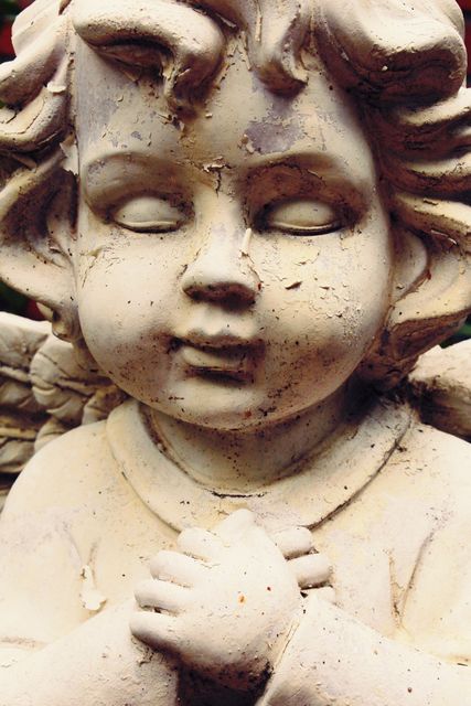 Weathered cherub angel statue capturing a moment of innocence with eyes closed and hands clasped in prayer. The pale surface shows signs of aging and distress, adding a rustic charm. Ideal for use in materials related to spirituality, religion, peace, reflection, or as decoration for gardens, memorials, and art projects.