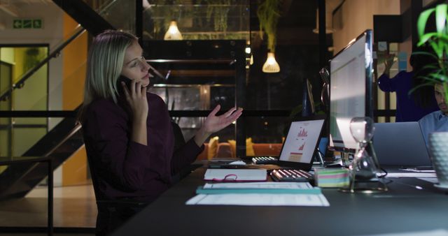 Businesswoman engaged in phone conversation while working on computer in modern office setting, ideal for illustrating contemporary office environments, business communications, remote meetings, and professional lifestyles.