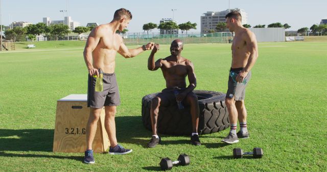 Three fit shirtless diverse men resting, drinking water and talking after exercising outdoors. cross training for fitness at a sports field.