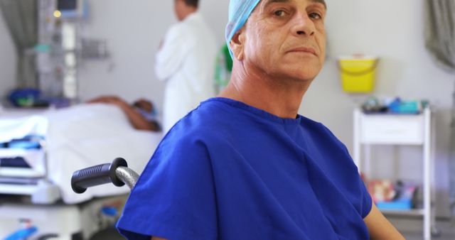 Senior man in a wheelchair wears a surgical gown and cap in a hospital ward. Background includes a medical professional attending to another patient and various medical equipment. Suitable for use in healthcare, elderly care, or patient treatment visuals.