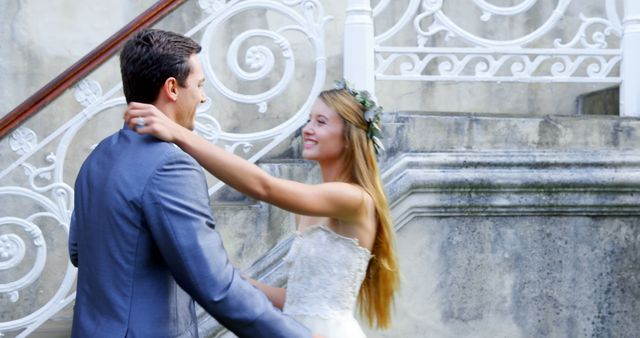 A young Caucasian bride and groom share a romantic moment on a staircase, with copy space. Their elegant attire and joyful expressions suggest a wedding celebration.