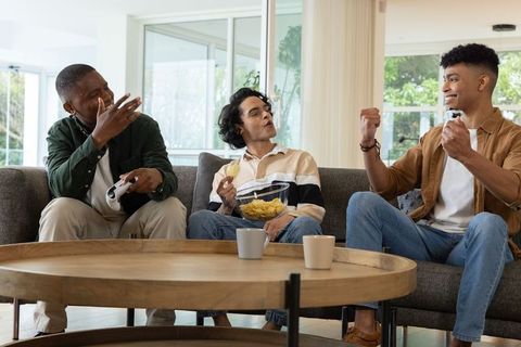 Diverse male friends playing video games on sofa while eating chips and laughing