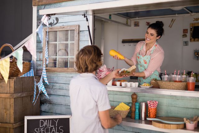 Waitress in apron serving a wrap to a customer at a food truck. The scene includes a chalkboard with daily specials, colorful drinks with straws, and a rustic wooden counter. Ideal for use in articles about street food, small businesses, customer service, and outdoor dining experiences.
