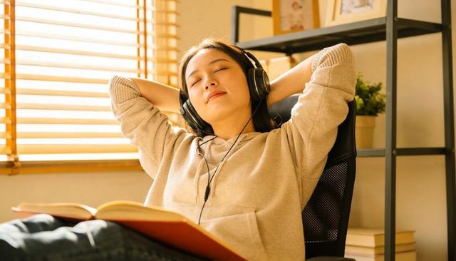 Asian woman relaxing in sunlight, listening with headphones and reading in cozy home study
