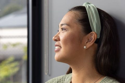 Mid-adult asian woman gazing out window with serene smile, headband and pearl earring