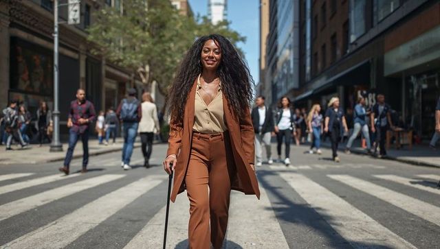 Confident professional walking with cane in rust tailored suit, beige blouse on city crosswalk