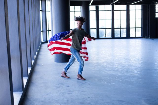 Transgender man holding American flag in empty parking garage, symbolizing freedom, identity, and diversity. Ideal for use in campaigns promoting gender expression, LGBTQ+ rights, and American pride. Suitable for articles, blogs, and social media posts focusing on modern urban life, inclusivity, and personal stories.