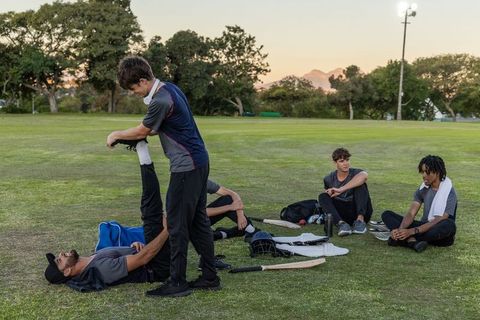 Diverse cricket teammates stretching on grassy field at dusk preparing for match