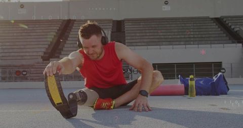 Male athlete stretching with prosthetic running blade on stadium track wearing red top and headphone