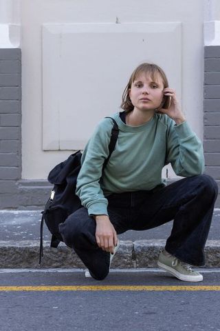 Contemplative young woman squatting on curb talking on phone with backpack, street style