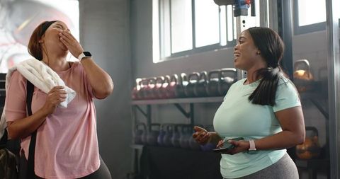 Diverse women laughing and chatting after workout by kettlebell rack in bright gym studio