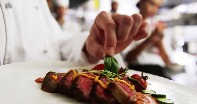 A chef is meticulously garnishing a beautifully cooked steak on a white plate. The background shows a busy professional kitchen with other chefs blurred in the distance, emphasizing the focus on the garnishing process. This image can be used for culinary blogs, restaurant promotions, or food magazine articles highlighting professional cooking techniques and fine dining experiences.