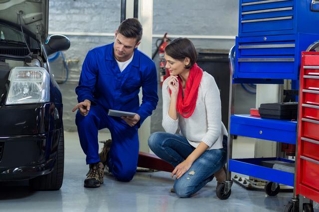 Mechanic in blue coveralls kneeling beside car, showing customer in white sweater and red scarf the issue with vehicle in repair garage. Useful for illustrating automotive services, customer service in repair shops, and professional car maintenance.