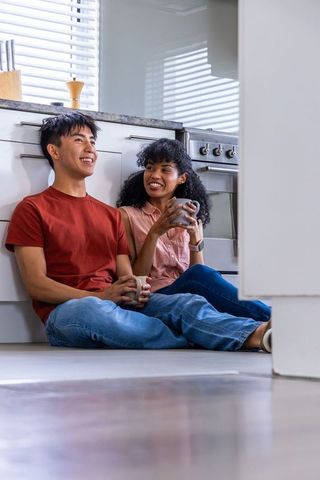 Couple sitting on kitchen floor sipping mugs with rising wellness icons and natural light