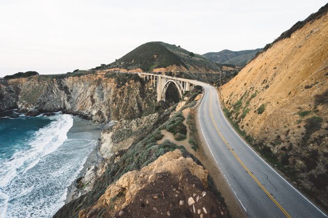 Photo shows a scenic coastal highway with a bridge winding through rocky cliffs along the seaside. Ideal for travel, adventure, road trip themes, or promoting scenic drives. Perfect for advertisements, travel blogs, magazines, or tourism websites focused on road trips and nature exploration.