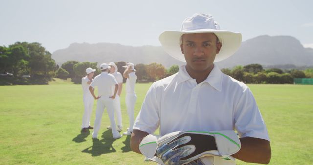 A young cricketer proudly standing in forefront wearing gloves and a hat with teammates chatting in background on a sunny cricket field. Ideal for promoting sports events, clubs, fitness programs, or teamwork-oriented campaigns. Suitable for articles, blogs, magazines focusing on cricket, sportsmanship, or youth activities.