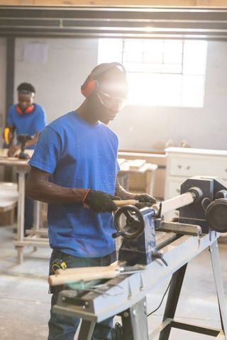 African american carpenter shaping wood on lathe in sunlit workshop with colleague drilling