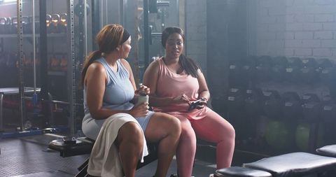 Plus-size women resting and chatting on gym bench after strength training session