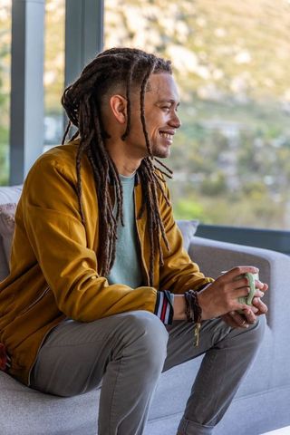 Smiling man with dreadlocks holding mug on sunlit sofa, casual relaxed morning at home