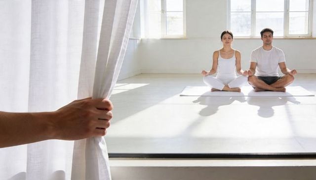 Hand pulling sheer curtain revealing couple meditating in sunlit minimalist yoga studio