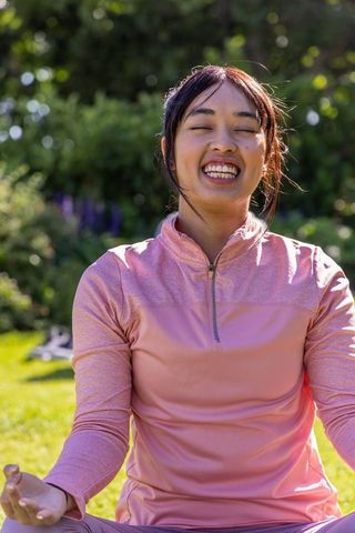 Smiling woman meditating on sunlit lawn wearing pink zip-up pullover for wellness