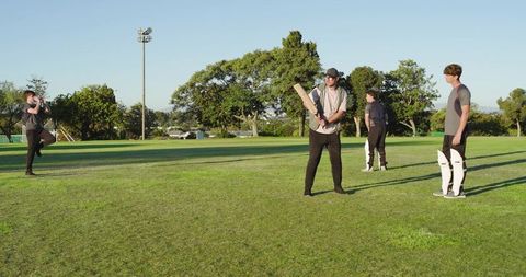 Male friends practicing cricket batting and fielding on sunny grass outfield
