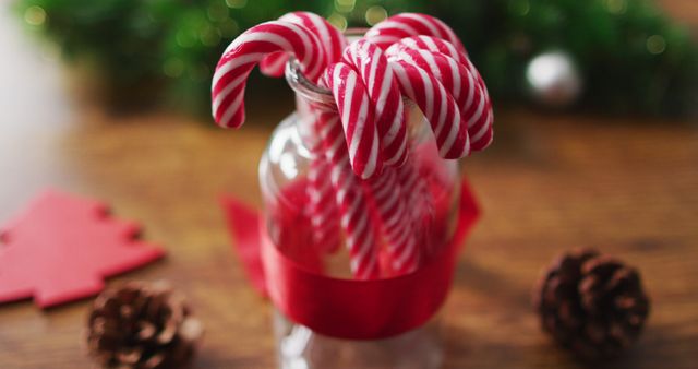 Candy canes in a glass jar wrapped with a red ribbon, with pine cones on a wooden table. Ideal for holiday greeting cards, festive promotional materials, and Christmas-themed social media posts. Great for celebrating the holiday season and adding a festive touch to any project.