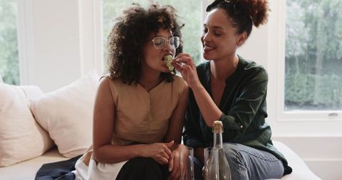 African American Female Couple Feeding Each Other Grapes on Cozy Sunlit Sofa