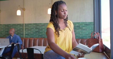 Woman reading paperback while reaching for coffee at window counter, brown handbag nearby