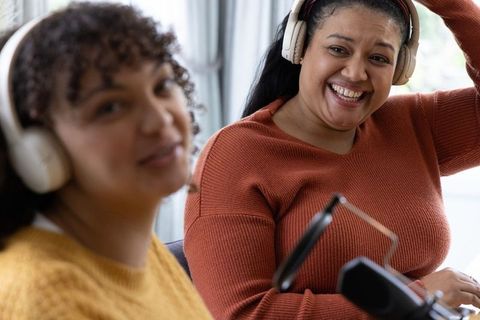 Two women recording podcast and laughing in cozy home studio wearing headsets