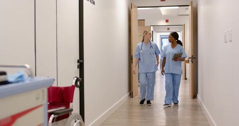 Two female nurses walking and talking in hospital corridor holding tablet and stethoscope