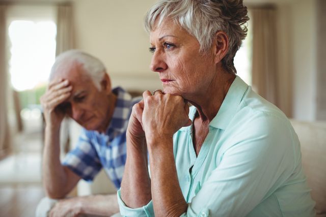 Senior couple sitting on sofa in living room, both looking upset and contemplative. Ideal for use in articles or advertisements about aging, relationships, mental health, and retirement challenges.