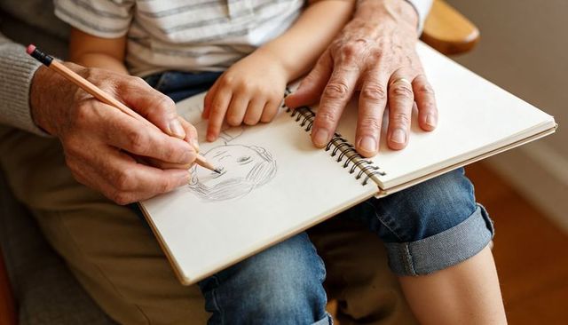 Grandparent guiding grandchild sketching portrait on lap with spiral sketchbook and pencil