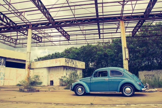 Classic blue Beetle parked at an old, abandoned gas station with a rusted overhead metal structure. The location features graffiti-covered walls and overgrown vegetation. Ideal for use in themes related to nostalgia, adventure, retro style, history, or automotive history.