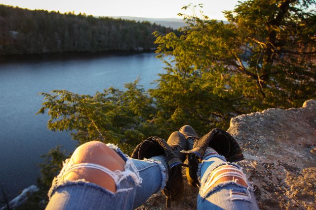 Person wearing ripped jeans and boots relaxing on a rocky overlook, enjoying a peaceful view of a forested lake at sunset. Ideal for use in travel blogs, nature explorations, leisure activities advertisements, and outdoor lifestyle promotions.