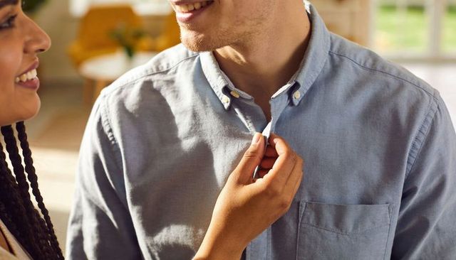 Woman buttoning man's light-blue button-down while couple sharing tender smile in sunlit living room
