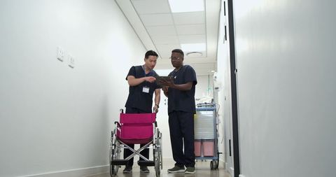 Asian and black nurses collaborating with tablet while checking pink wheelchair in hospital
