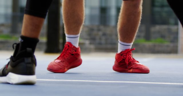 Feet of basketball players in athletic shoes on an outdoor court, one wearing red shoes and the other black. Suitable for use in articles, advertisements, or media related to sports, fitness, athletic footwear, basketball tips, and outdoor activities.